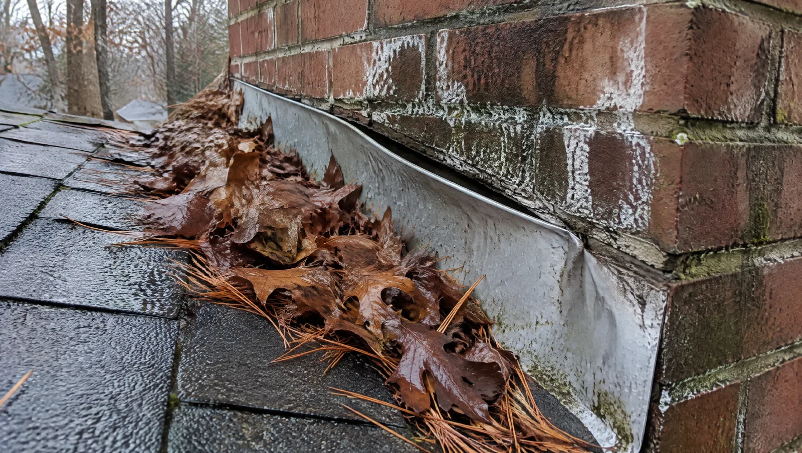 Wet leaves piled against lifted flashing on the uphill side of a brick chimney