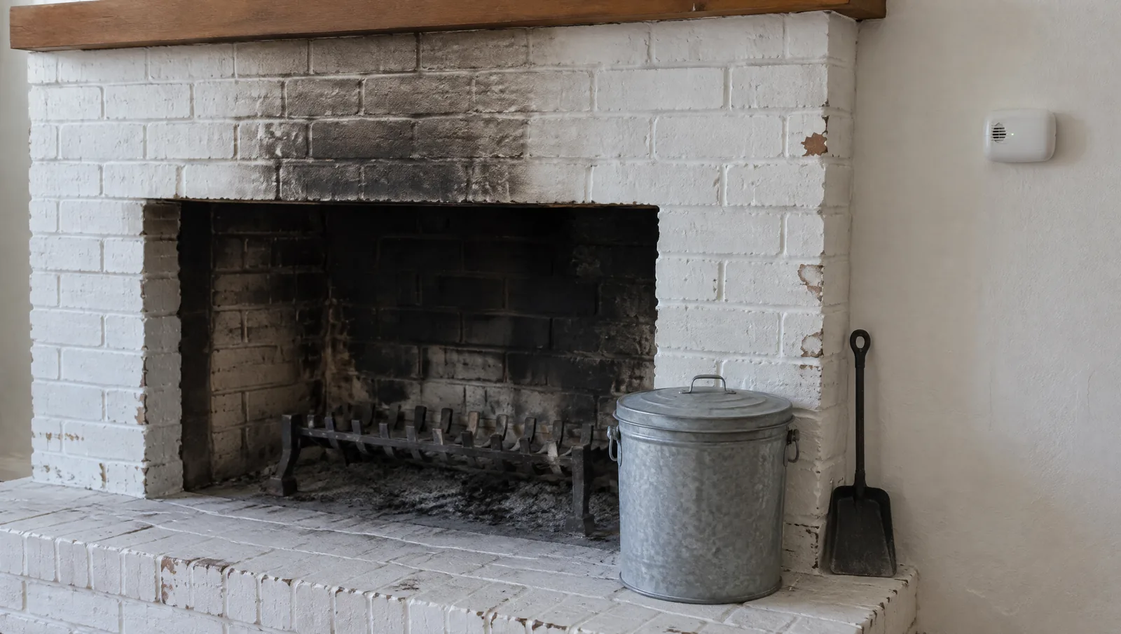 Painted brick fireplace with a lidded metal ash can on the hearth and a carbon monoxide alarm beside it