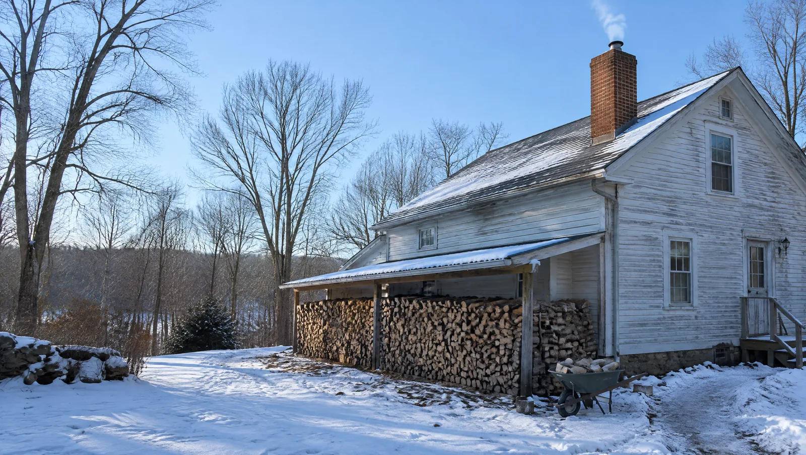 Farmhouse chimney with a thin wisp of wood smoke and a long stack of firewood along the house in snow